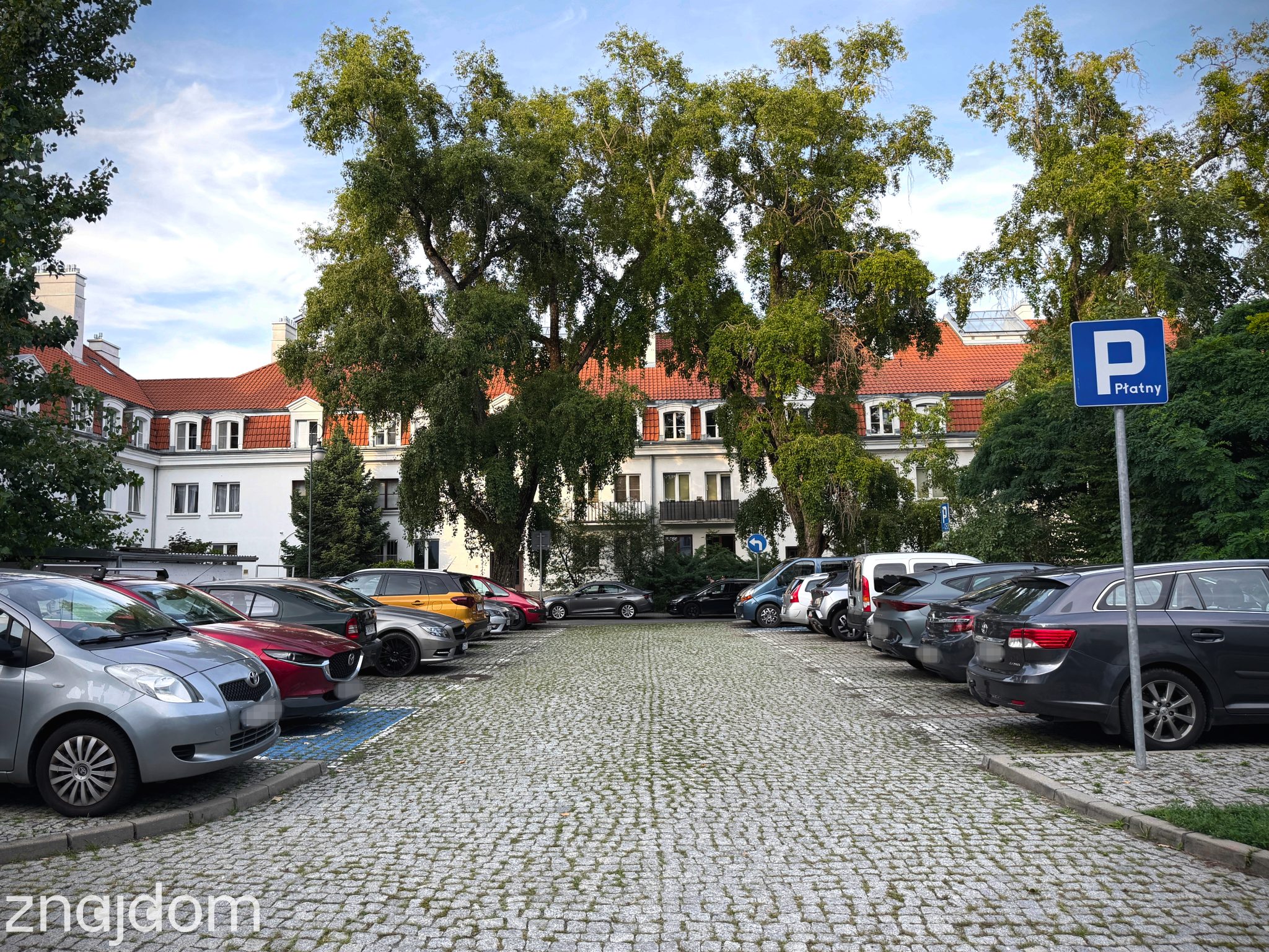 Pełny parking pod fasadą modernistycznego osiedla mieszkaniowego.
