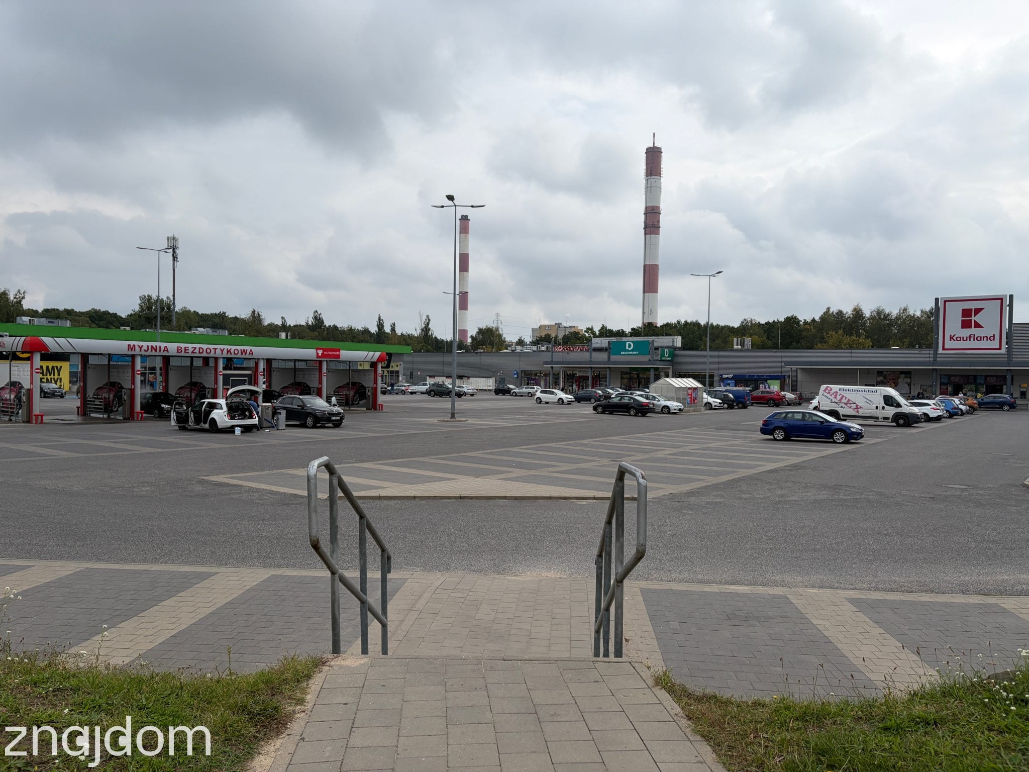 Widok ze schodków na parking centrum handlowego przy Rondzie Sybiraków. Na parkingu myjnia.