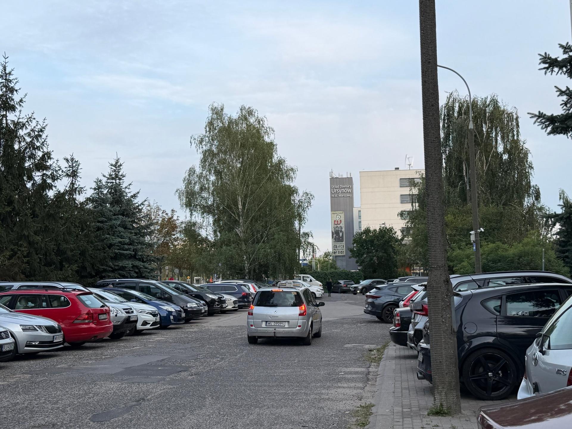Gęsto zastawiony samochodami, ciasny parking na warszawskim Ursynowie z widocznym w tle budynkiem Urzędu Dzielnicy.