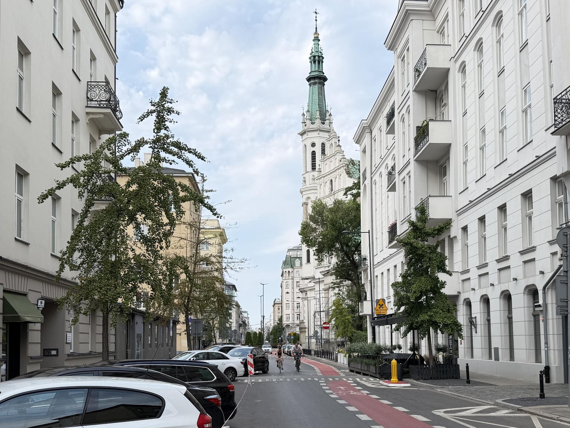 Śródmiejska ulica z czerwoną ścieżką rowerową, eleganckimi kamienicami po bokach i wysoką wieżą kościoła w tle w Warszawie.