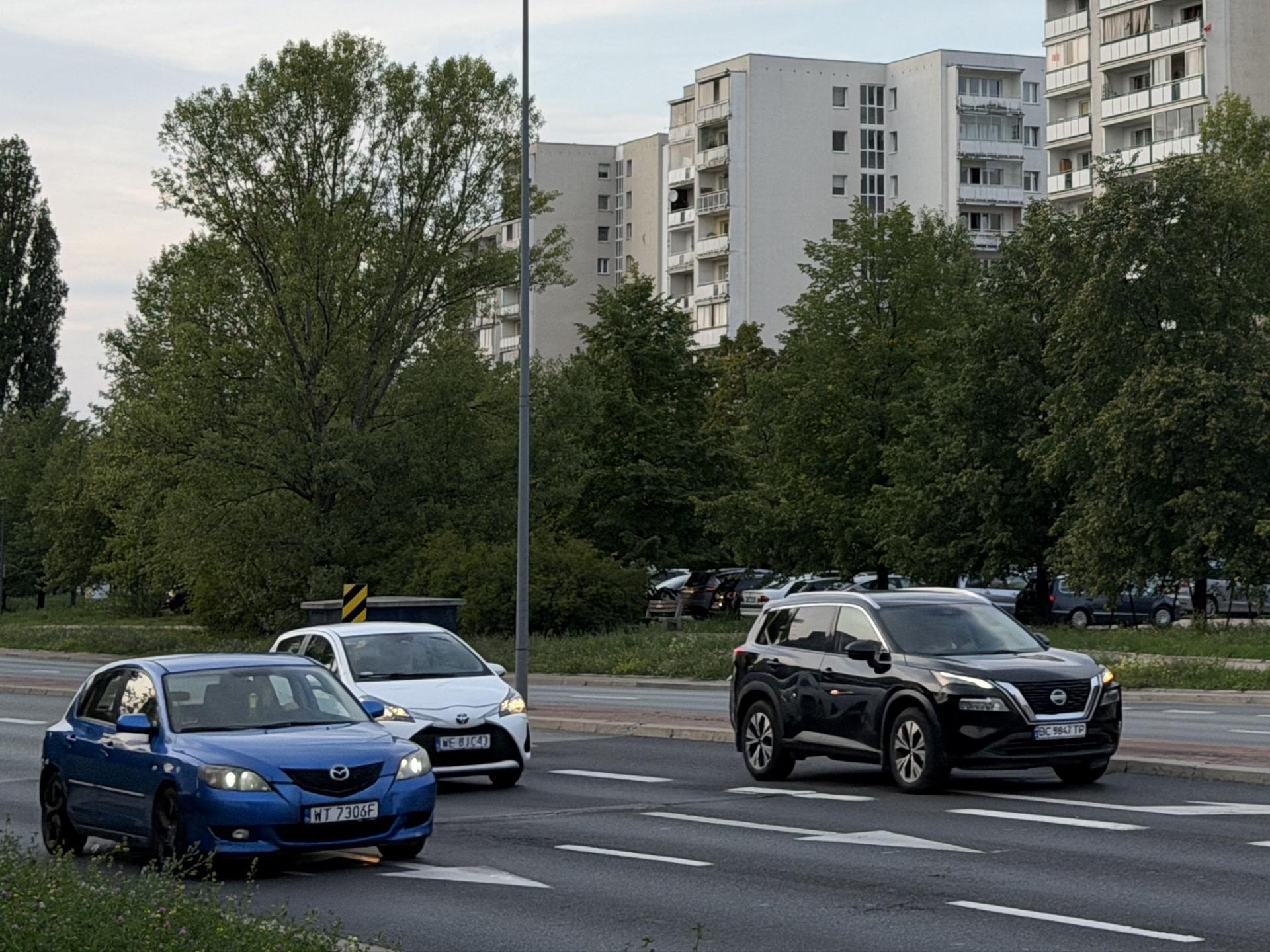 Ruch samochodowy na wielopasmowej ulicy na tle drzew i wysokiego bloku mieszkalnego na Ursynowie.