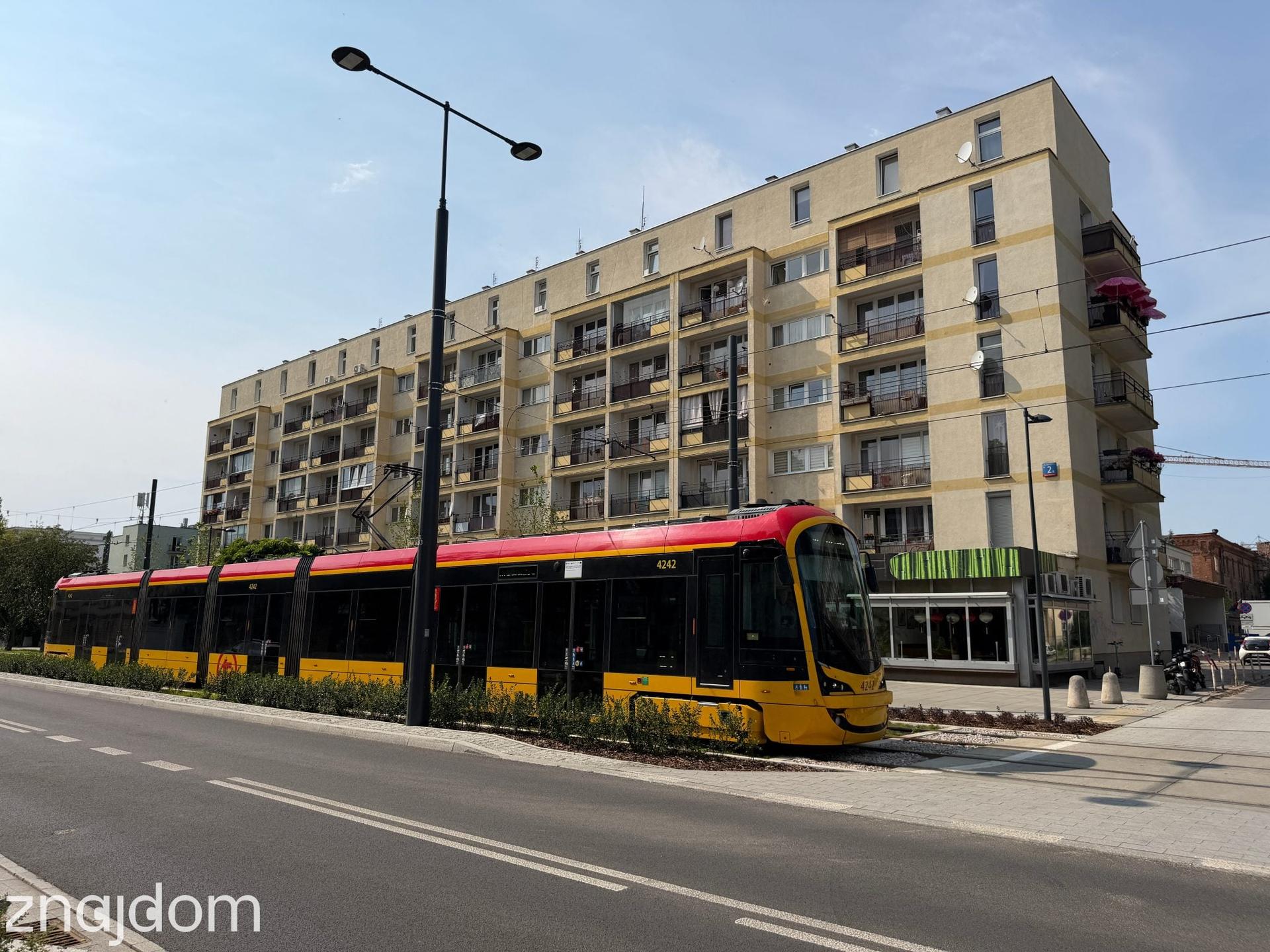 Nowoczesny, niskopodłogowy warszawski tramwaj jadący wzdłuż wielorodzinnego bloku mieszkalnego na Mokotowie.