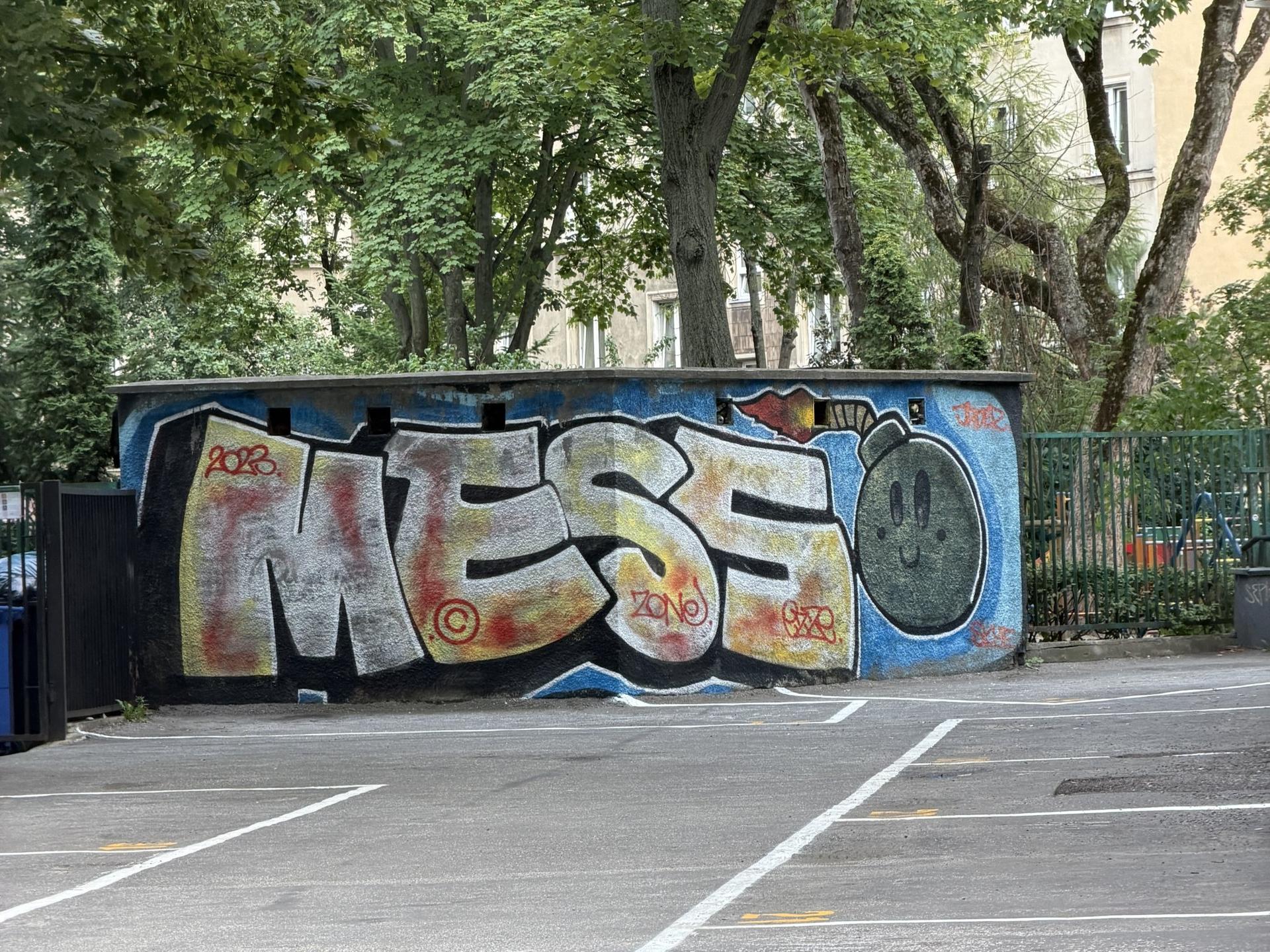 Kolorowe graffiti z dużym napisem i rysunkiem na betonowym murku, w otoczeniu zieleni na warszawskim Śródmieściu.
