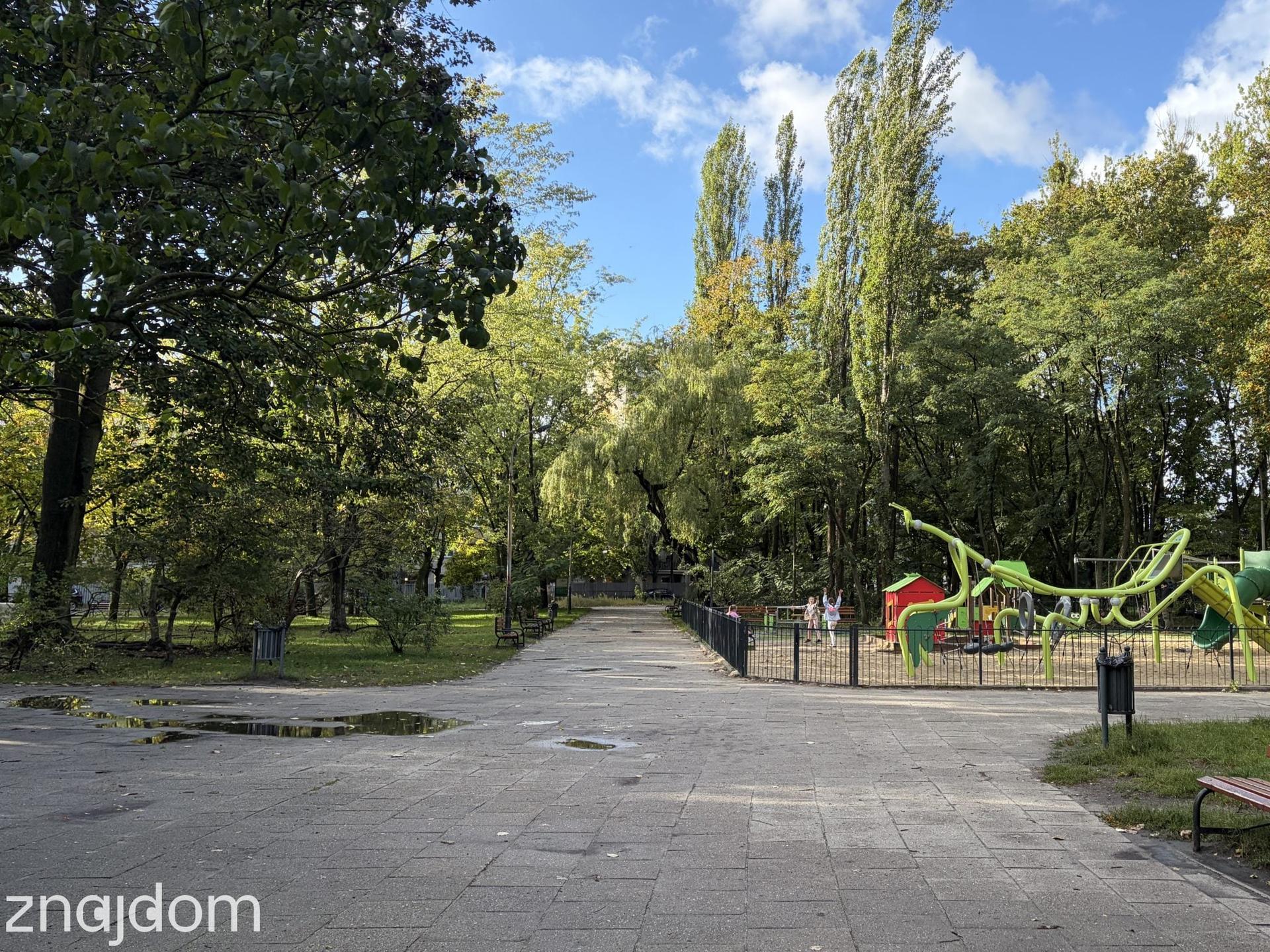 Szeroka aleja spacerowa otoczona wysokimi drzewami, obok której znajduje się ogrodzony, zielony plac zabaw.