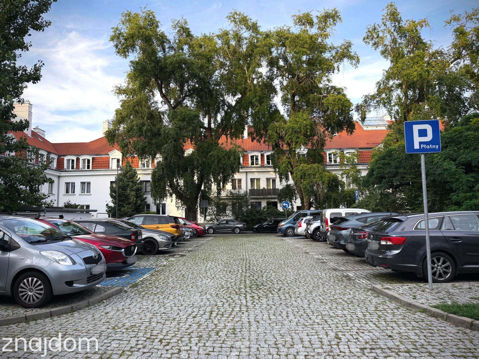 Pełny parking pod fasadą modernistycznego osiedla mieszkaniowego.