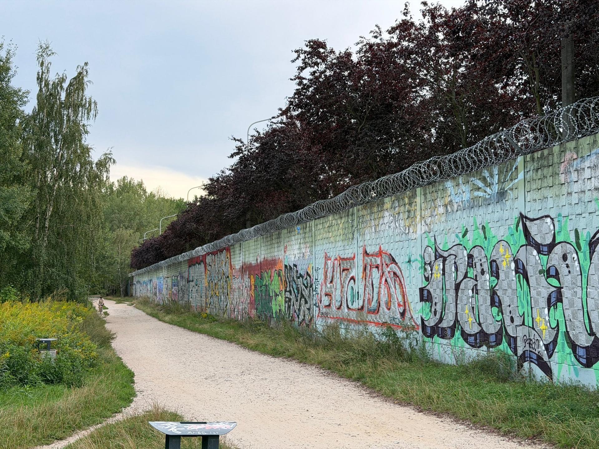 Długi mur pokryty graffiti i zwieńczony drutem kolczastym, ciągnący się wzdłuż piaszczystej ścieżki spacerowej na warszawskim Ursynowie.