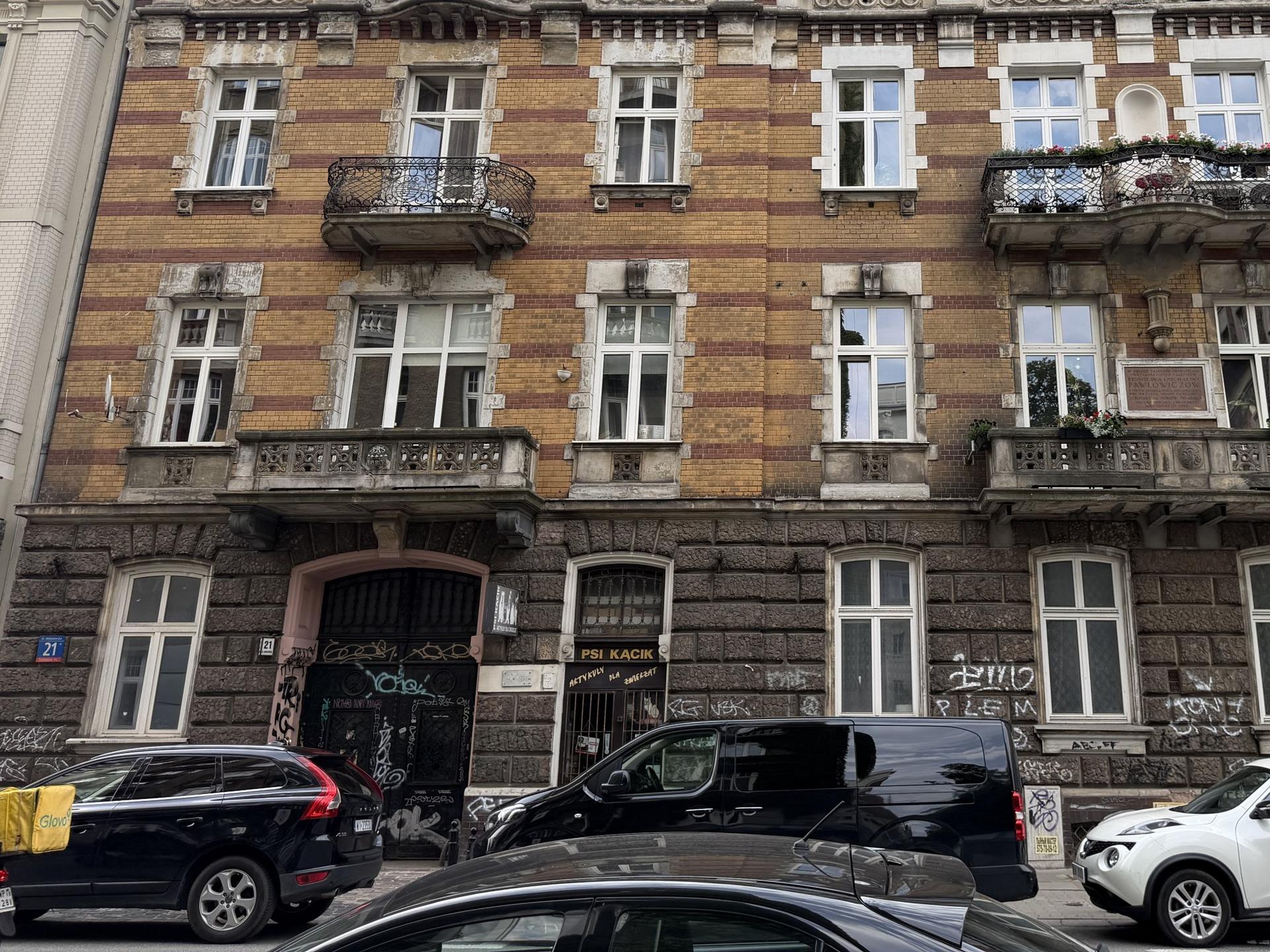Zabytkowa, wielopiętrowa kamienica z widocznymi ubytkami w elewacji i gęsto pomazanym graffiti parterem oraz zaparkowanymi autami na warszawskim Śródmieściu.