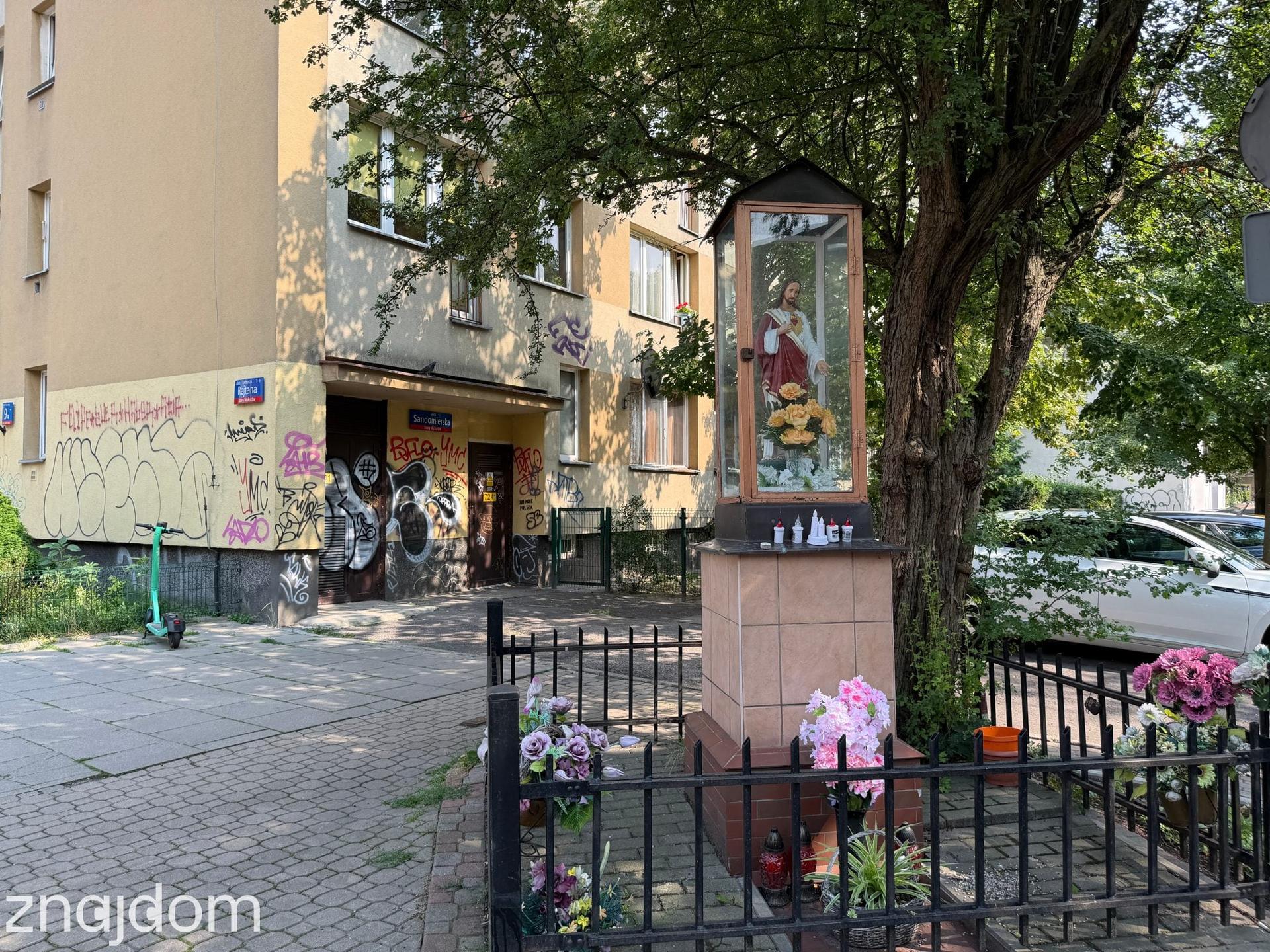 Zadbana kapliczka z figurą i kwiatami na tle starszego budynku mieszkalnego z elewacją zniszczoną przez graffiti na Mokotowie.