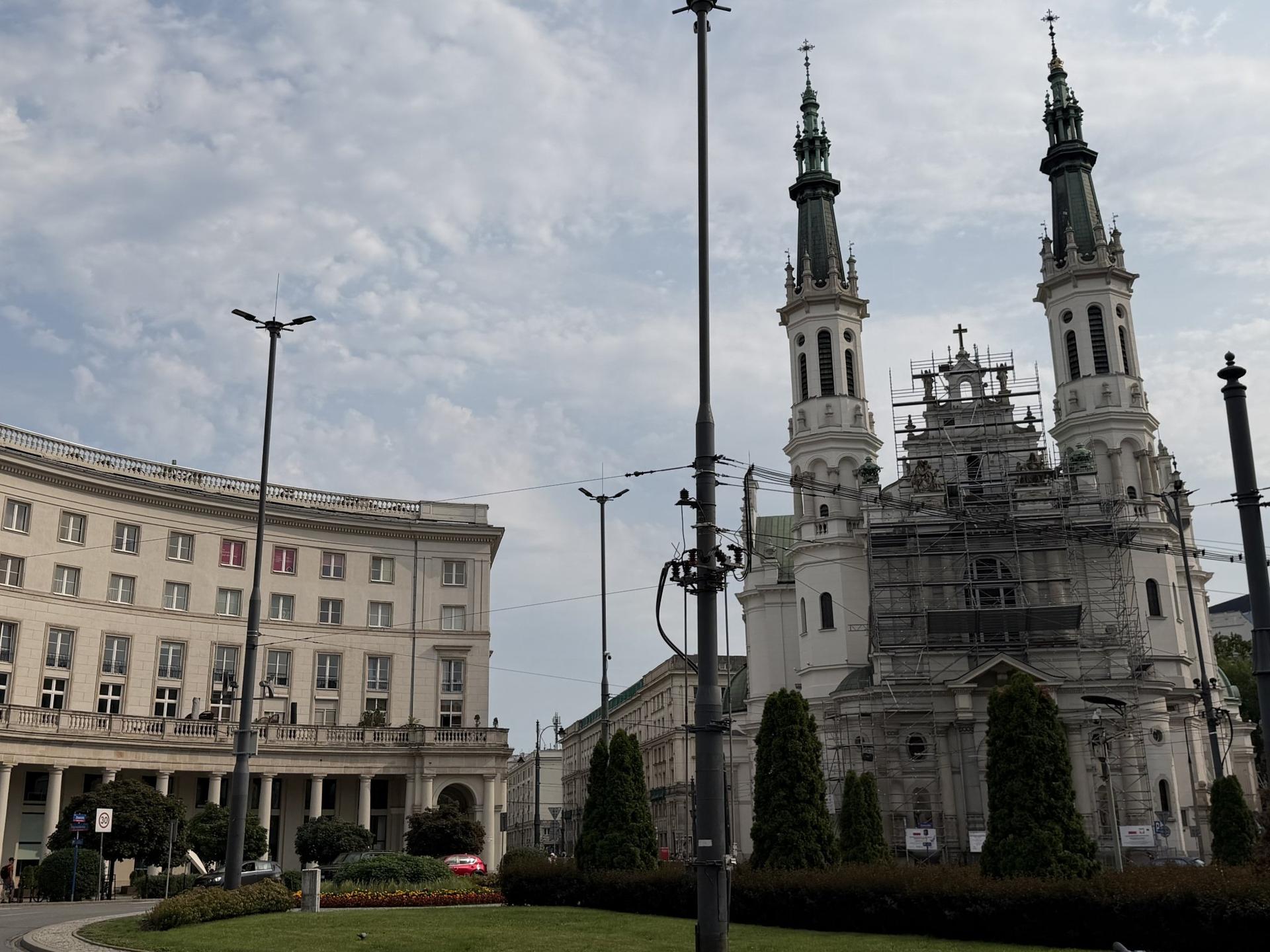 Widok na Plac Zbawiciela w Warszawie z charakterystycznymi arkadami budynków i strzelistymi wieżami kościoła na tle jasnego nieba.
