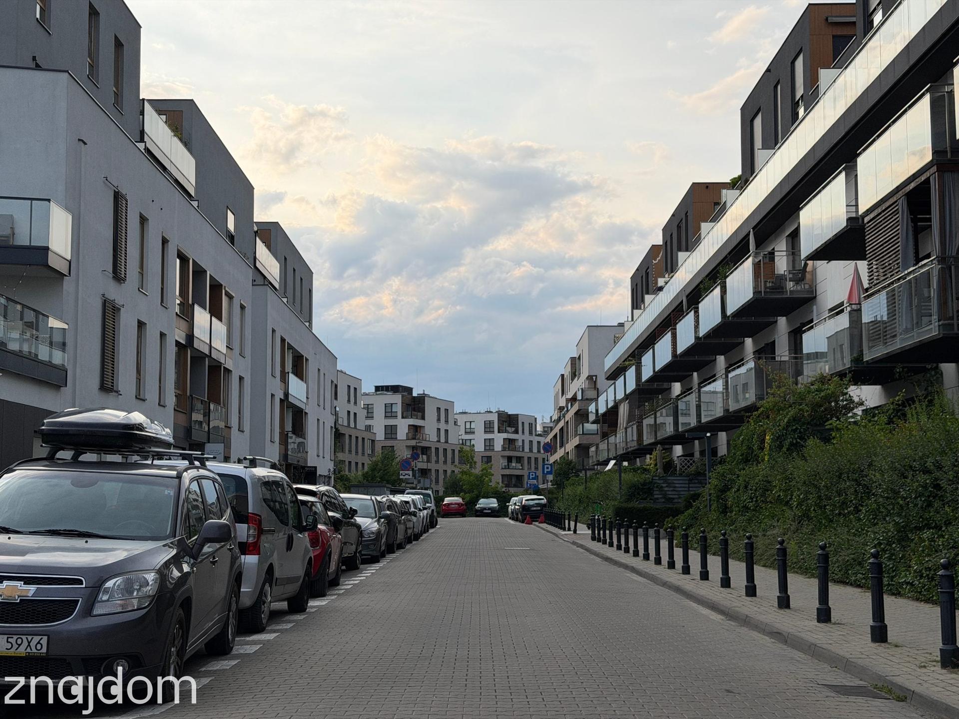 Widok na uliczkę między apartamentowcami na Wilanowie. Wszystkie miejsca parkingowe są zajęte.