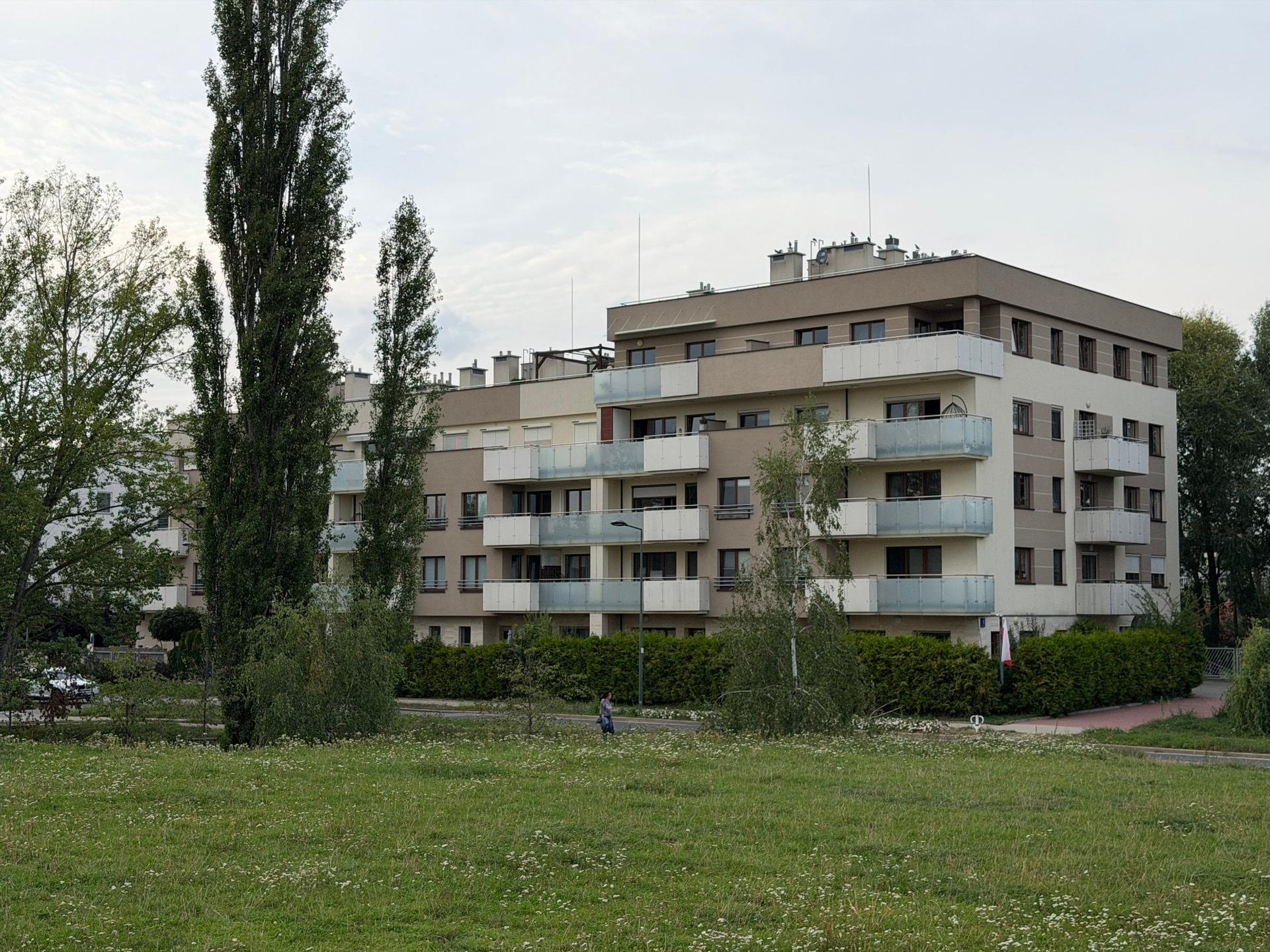 Nowoczesny, niski apartamentowiec z przeszklonymi balkonami, oddzielony od ulicy szerokim pasem zieleni i wysokimi drzewami na warszawskim Ursynowie.