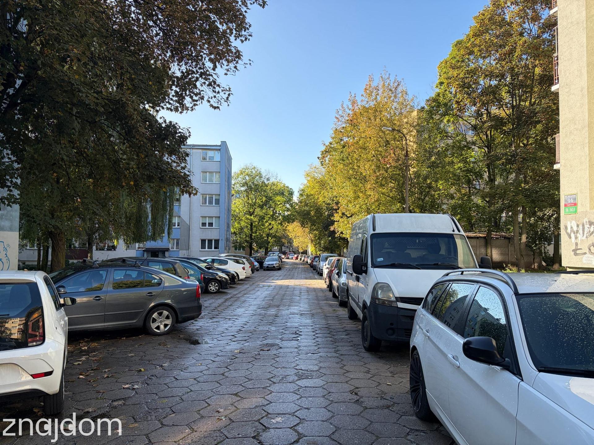 Wąska, osiedlowa ulica gęsto zastawiona zaparkowanymi samochodami na tle bloku mieszkalnego i wysokich drzew.