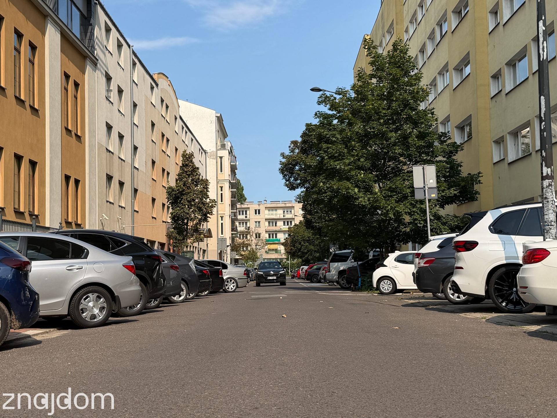 Sznur gęsto zaparkowanych samochodów zajmujących chodniki po obu stronach ulicy ze starszą zabudową wielorodzinną na Mokotowie.