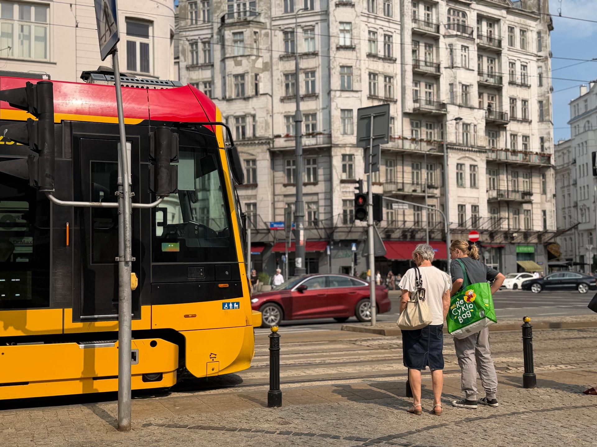 Żółto-czerwony tramwaj na przystanku przy przejściu dla pieszych, na tle zabytkowych kamienic i ruchu ulicznego w warszawskim Śródmieściu.