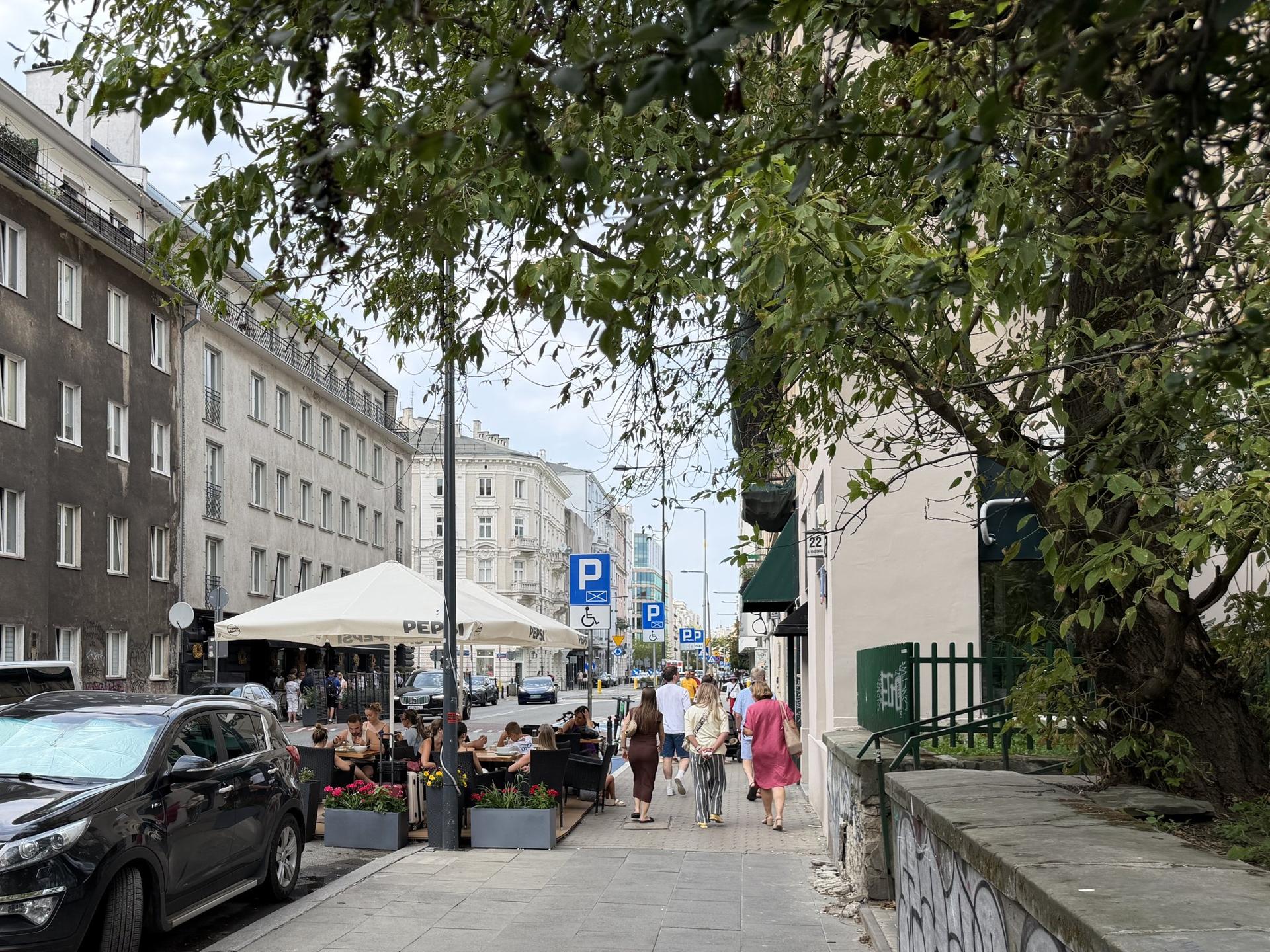 Spacerujący przechodnie i goście w letnim ogródku restauracyjnym na gwarnej ulicy warszawskiego Śródmieścia w otoczeniu zabytkowych kamienic.