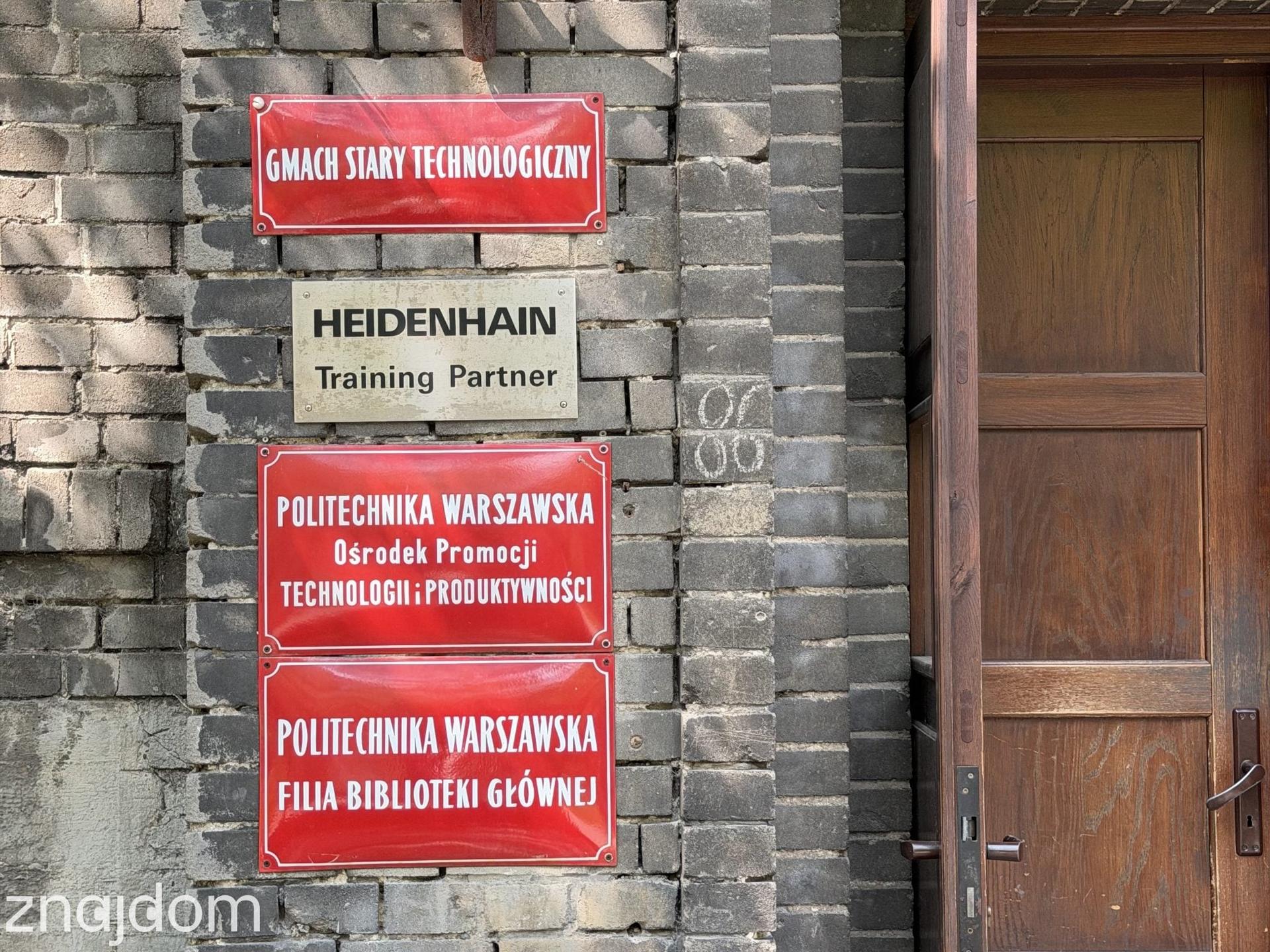 Ceglany fragment elewacji z drewnianymi drzwiami i czerwonymi szyldami informacyjnymi Politechniki Warszawskiej na Mokotowie.