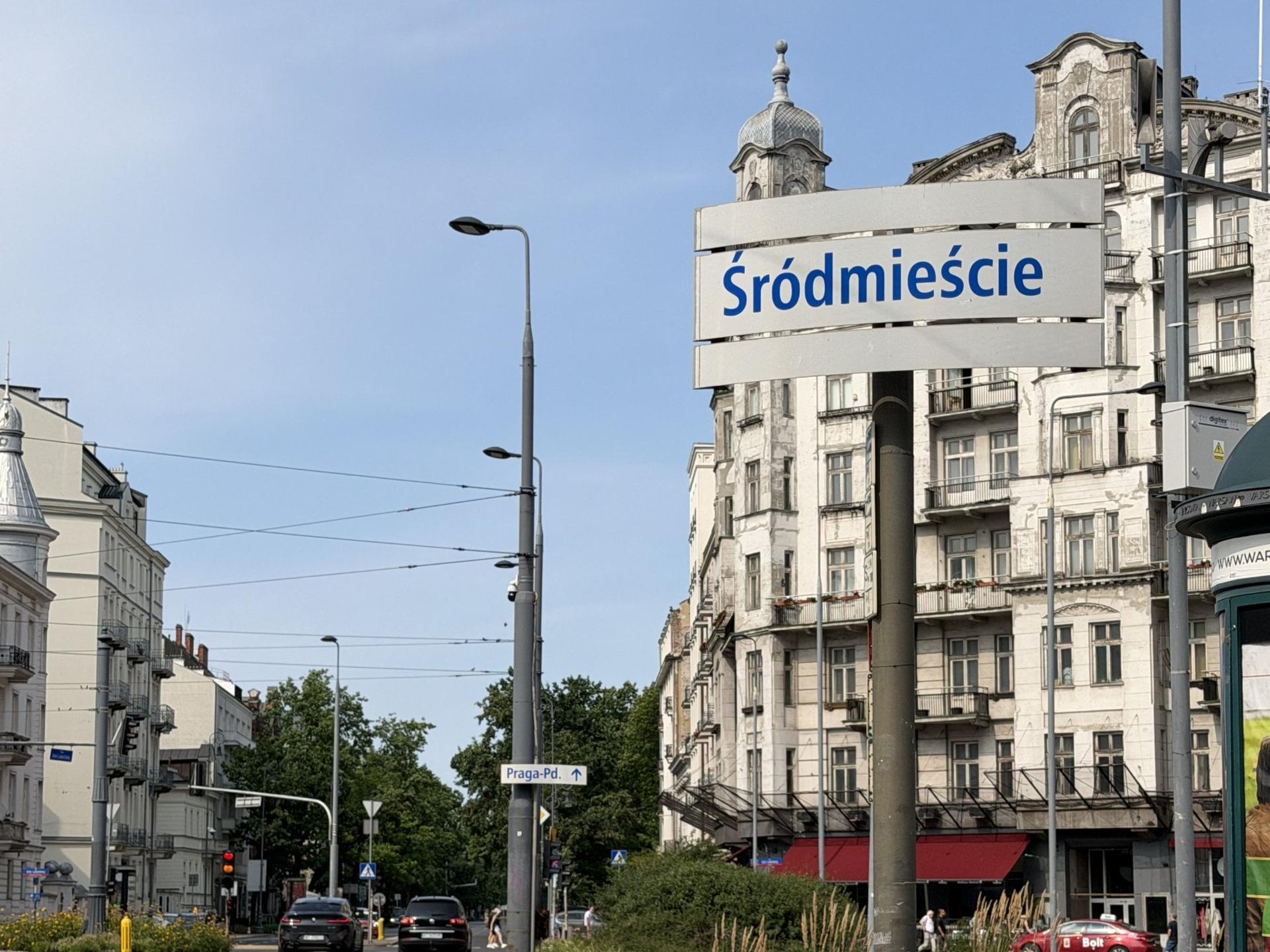 Tablica informacyjna z napisem „Śródmieście” na tle wysokich, zabytkowych kamienic i miejskiego ruchu ulicznego w Warszawie.