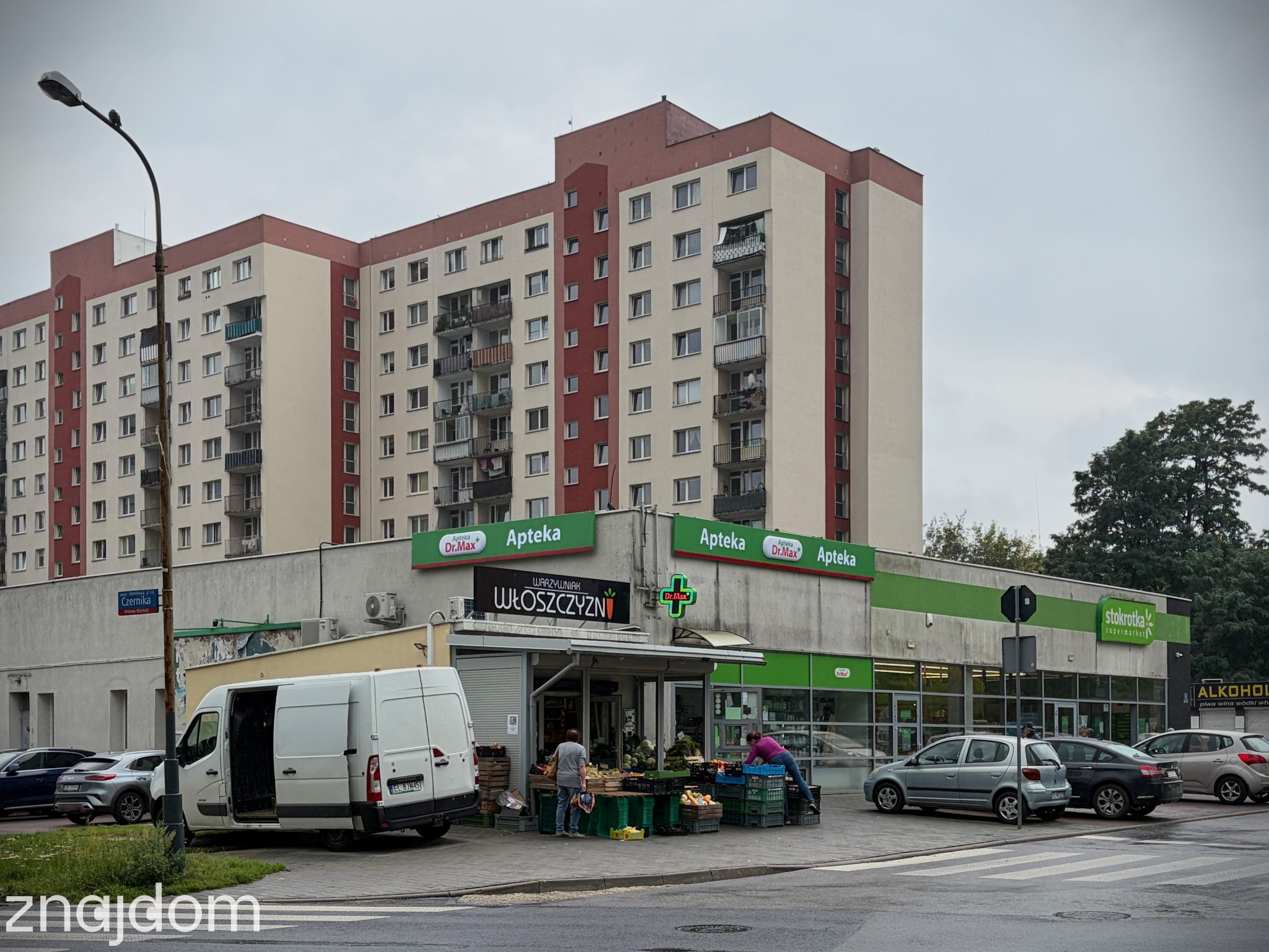 Zdjęcie budynku usługowego w ciągu dnia. Widać aptekę i superkmarket. W tle wysokie bloki.